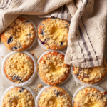 Nio blåbärs- och vaniljmuffins med smuldeg på en plåt, delvis täckta av en kökshandduk